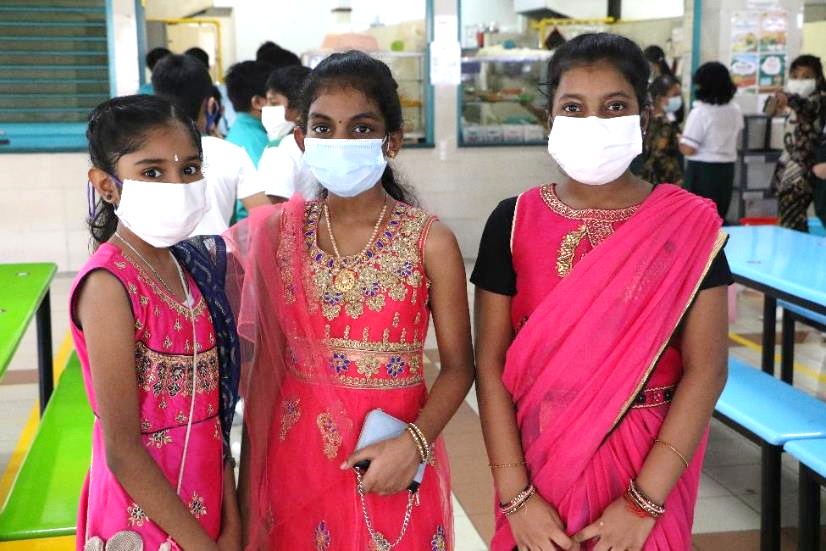 Three girls in colorful traditional Indian attire wearing white face masks stand together indoors.